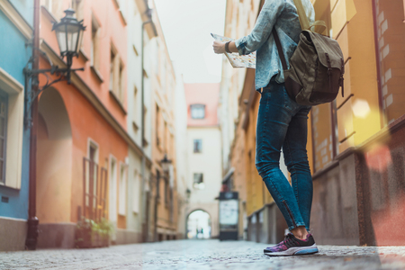 Young european female tourist with a backpack standing on a narrow street in the old town holding map with city sights and looking for the next place to go. View from the back. Copy space for your text.の写真素材
