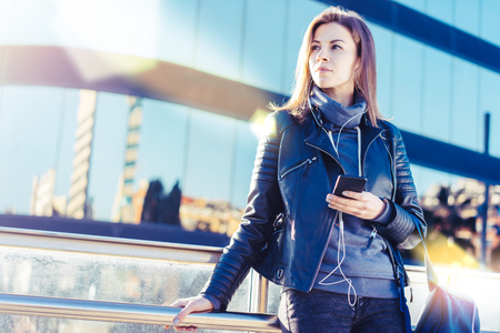 Portrait of cute backpacker girl in leather biker jacket looking aside while listening to music on her mobile phone. Contemporary business centre on the background. Copy space for your text.の写真素材