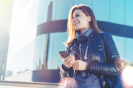 Portrait of smiling young woman in leather black  jacket looking aside while listening to music on her mobile phone. Female backpacker being happy enjoying beautiful sunny day-off. Contemporary business centre on the background. Copy space for your text.の写真素材