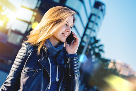 Portrait of attractive young woman in leather biker jacket talking on her mobile phone standing in front of modern hi-tech style glass building of business centre. Blurred background. Copy space for your text.の写真素材