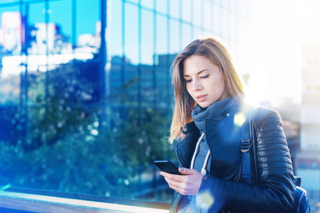 Attractive young woman in leather biker jacket reading message on her mobile phone standing in front of modern hi-tech style glass building of business centre. Blurred background. Copy space for your text.の写真素材