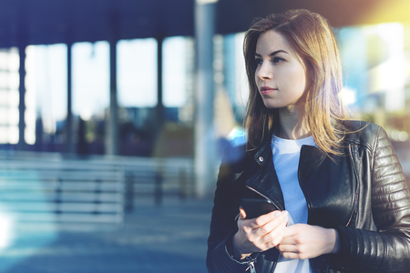 Portrait of attractive student girl having day off spending it outdoors, looking aside with mobile phone in her hands. Blurred background. Copy space for your text.の写真素材
