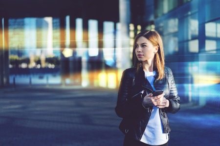 Portrait of attractive young woman in black leather biker jacket and white basic t-shirt standing on a street in front of shopping mall, holding mobile phone in her hands and looking aside. Blurred background. Copy space for your text.の写真素材