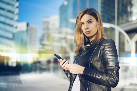 Portrait of cute girl in leather biker jacket holding in hands mobile phone and looking to the side while sending message. Modern, hi-tech style building on the background. Copy space for your text.の写真素材