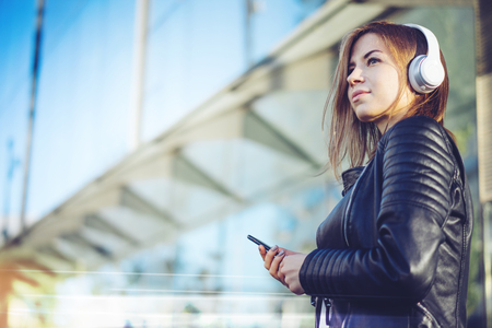 Close up portrait of young cute girl wearing black leather biker jacket looking aside while listening to music on her mobile phone and enjoying sunny day off. Modern glass building of business centre on the background. Copy space for your text.の写真素材