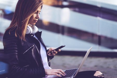 Attractive freelancer girl dressed in black leather biker jacket and basic white t-shirt working on her laptop and reading message on mobile phone while sitting outdoors on a bench in front of modern glass building of business centre. Copy space for your text.の写真素材