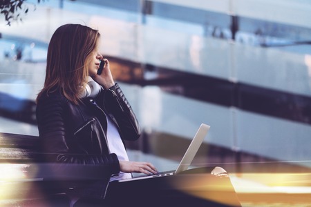 Stylish freelancer girl dressed in black leather biker jacket and basic white t-shirt working on her laptop and talking on mobile phone while sitting outdoors on a bench in front of modern glass building of business centre. Copy space for your text.の写真素材