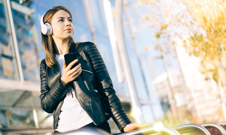 Portrait of attractive girl in leather biker jacket enjoying listening to music on her mobile phone while having rest outdoors sitting in front of modern business centre. Urban lifestyle concept.の写真素材