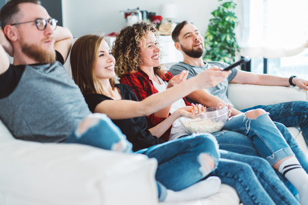 Side view of four friends sitting on sofa and watching tv in cozy light colored living room. Two...