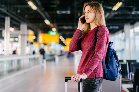 Female passenger standing with her bag and backpack and talking on mobile phone while waiting for delayed flight in the international airport terminal. Blurred background. Focus is on the hand holding case handle.の写真素材