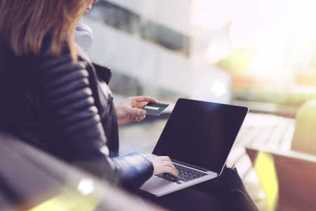 Cropped photo of a girl in black biker leather jacket sitting on a bench outdoors with her laptop, holding credit card in hand and making online purchases. Internet shopping concept. Close up. View from the back.の写真素材