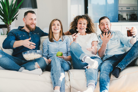Group of young people having day-off spending their time on sofa in front of tv in cozy living room. Four best friends being very excited celebrating their favourite team victory after watching football mach.の写真素材