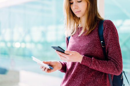Close up photo of female passenger standing near the window with passport and boarding pass in her hand, checking message on her mobile phone while waiting for delayed flight in the international airport terminal. Cropped shot with blurred background.の写真素材