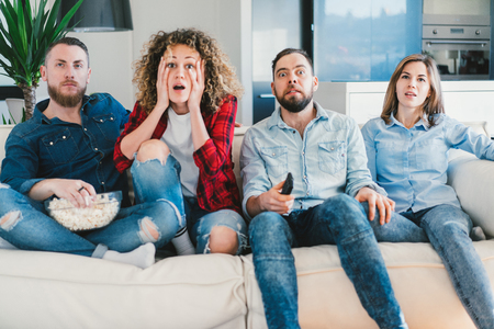 Side view of four friends sitting on sofa and watching football world cup in cozy light colored apartment. Young people cheering and being excited about the score.の写真素材