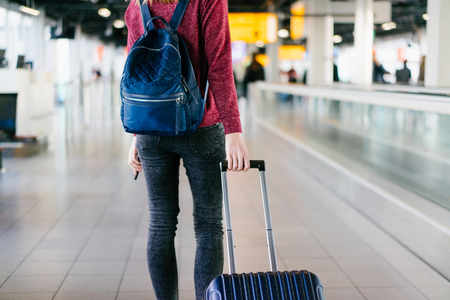Cropped photo of female passenger with bag and backpack walking through the international airport terminal looking for her gate. Blurred background. View from the back.の写真素材