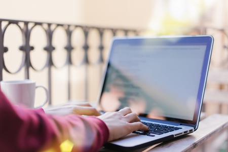 Close up cropped shot of female freelancer sitting on sunlight filled terrace and working on her laptop. Woman hands typing on a keyboard. View from the back. Horizontal.の写真素材