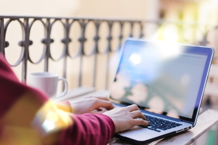 Close up cropped shot of female freelancer sitting on sunlight filled terrace and working on her laptop. Woman hands typing on a keyboard. View from the back. Horizontal.の写真素材