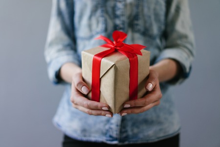 Close up cropped shot of a woman dressed in denim shirt  holding gift box wrapped in craft paper and decorated with red satin ribbon in her hands. Plain grey background.の写真素材