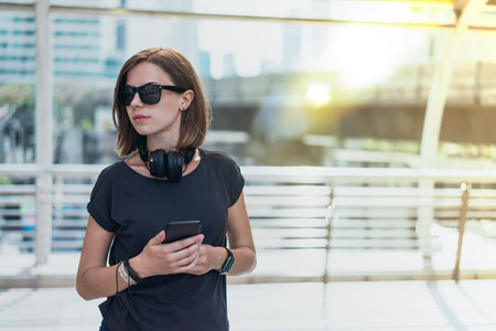 Young female hipster standing at the city rail station holding in hands her mobile phone while waiting for the train. Mock up, copy space for your text. Urban life concept. Copy space for your text.の写真素材