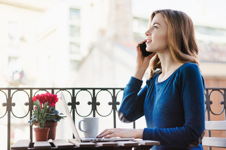 Young smiling attractive casual dressed business woman sitting on terrace working on her laptop and talking on mobile phone discussing new project with her partners. Distant job in comfortable conditions concept. Side view.の写真素材
