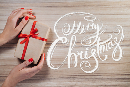 Holiday composition. Cropped shot of female hands holding gift wrapped in craft paper and decorated with red satin ribbon. Merry Christmas wishes written with calligraphic font. Top view, flat lay.の写真素材