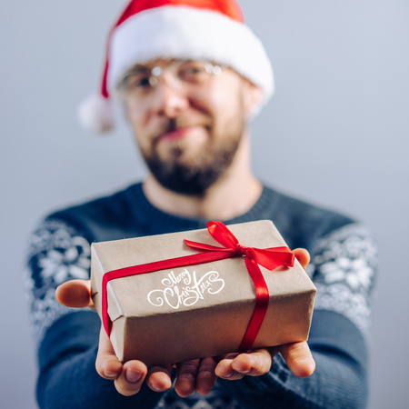 Front view. Christmas concept. Cropped shot of a bearded guy in santa clause hat and eyeglasses holding gift wrapped in craft paper and decorated with red sating ribbon in his hands.の写真素材