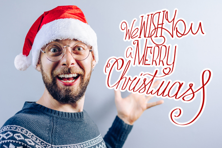 Front view. Christmas concept. Portrait of smiling bearded guy wearing santa hat, eyeglasses and blue knitted sweater with blank grey wall on the background. Handdrawn x-mas wishes.の写真素材