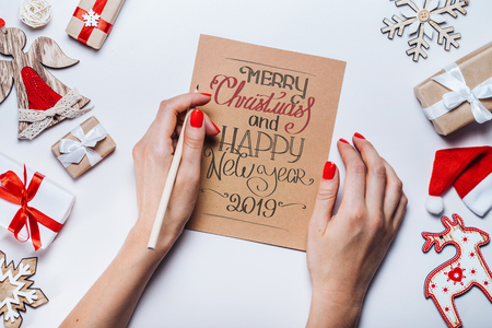Christmas decorations and gift box on wooden desk. Female hands holding pencil writing best wishes on the   piece of craft paper. Top view, flat lay.の写真素材