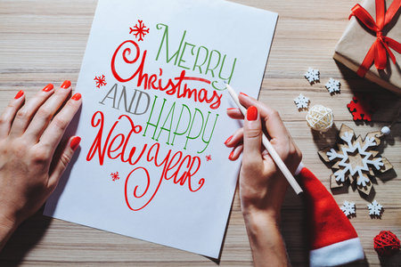 Christmas decorations and gift box on wooden desk. Female hands holding pencil writing best x-mas...