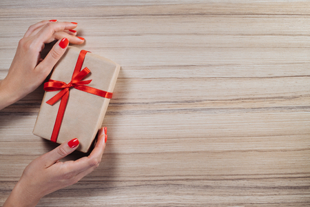 Christmas composition. Top view, flat lay. Female hands holding gift box wrapped in craft paper and decorated with red satin ribbon on wooden background. Copy space for your text.の写真素材