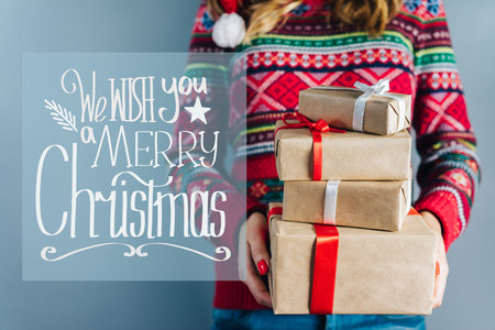 Blonde girl in red knitted sweater with christmas pattern holding bunch of gift boxes wrapped in craft paper and decorated with satin ribbon. Hand drawn x-mas wishes.の写真素材