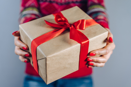 Cropped shot of girl in knitted sweater with traditional christmas pattern holding gift box wrapped in craft paper and decorated with red satin ribbon. New year concept.の写真素材