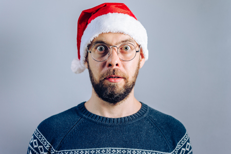 Front view. Christmas concept. Portrait of a bearded guy in santa hat and eyeglasses looking astonished being surprized by the great news.の写真素材