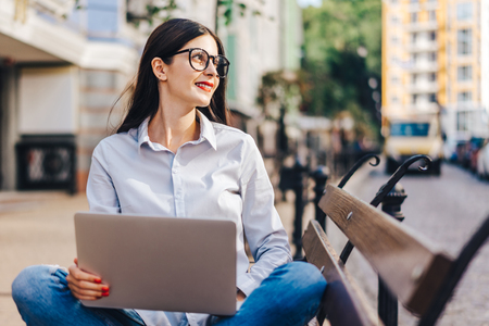 Pretty smiling casually dressed student girl sitting outdoors on a bank working using her laptop. Freelance, distant work and education concept.の写真素材