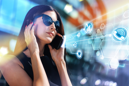 Attractive smiling brunette business woman with red lips wearing elegant black dress and sunglasses standing in front of hi-tech glass building of business center talking on her mobile  phone discussing new project with clients. Abstract background with roaming, lte, mobile applications infographics.の写真素材