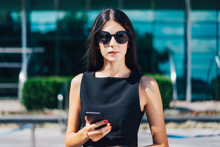 Beautiful brunette business woman wearing elegant black dress, sunglasses, with red lips and nails posing in front of hi-tech glass building of business center with mobile phone in hands. Front view close up cropped horizontal portrait.の写真素材
