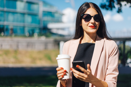 Beautiful smiling brunette business woman wearing elegant black dress, sunglasses, with red lips and nails having coffee break outdoors. Modern urban lifestyle concept. Copy space for your text.の写真素材