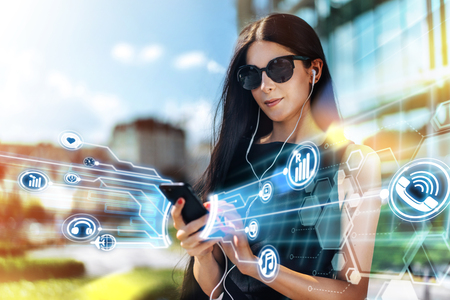 Beautiful brunette business woman wearing basic black dress, sunglasses standing near hi-tech glass building of business center with mobile phone in hands and listening to music. Modern urban lifestyle concept. Mobile applicatios, media and social network inforgraphics.の写真素材