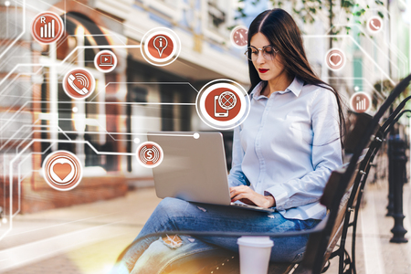 Young pretty female freelancer working outdoors using her laptop and mobile phone sitting on the street bench. Social network and mobile applications infographics.  Business, education and freelance concept.の写真素材