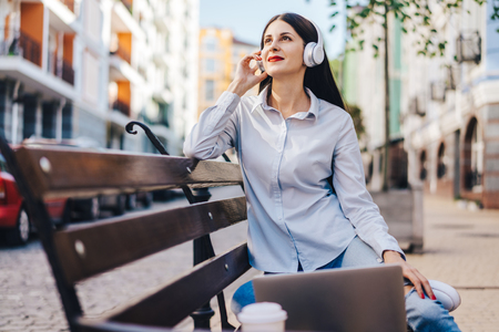 Pretty smiling casually dressed student sitting outdoors on a bank working on her laptop. Beautiful young european freelancer having coffee break and enjoyng music using generic design earphones. Freelance, distant work and education concept.の写真素材