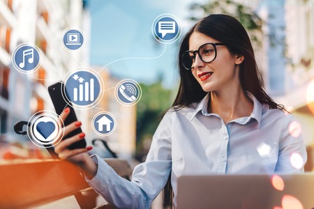 Young pretty female freelancer working outdoors using her laptop and mobile phone sitting on the street bench. Social network and mobile applications infographics.  Business, education and freelance concept.の写真素材