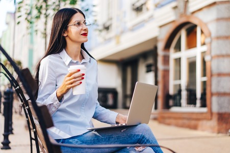 Pretty smiling casually dressed student girl sitting outdoors on a bank enjoyng her coffee and working using laptop. Freelance, distant work and education concept. Copy space for your text.の写真素材