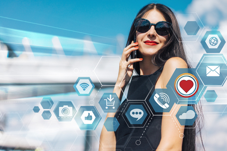 Attractive smiling brunette business woman with red lips wearing elegant black dress and sunglasses standing in front of hi-tech glass building of business center talking on her mobile  phone discussing new project with clients. Social network and mobile applications infographics.の写真素材