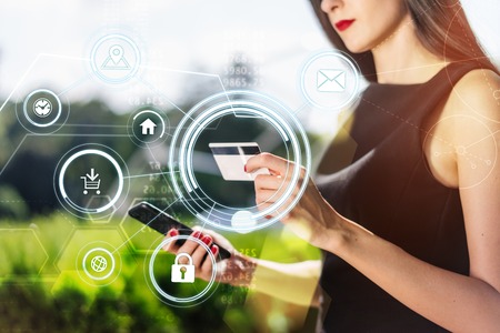 Cropped portpait of pretty brunette business woman wearing smart black dress standing  outdoors in front of hi-tech glass building of shopping mall with credit card in her hand using mobile phone for online purchases. Mobile applications infographics.の写真素材