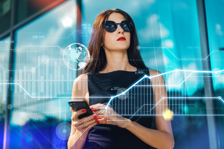 Beautiful brunette business woman wearing elegant black dress, sunglasses, with red lips and nails posing in front of hi-tech glass building of business center with mobile phone in hands. Abstract futuristic background with global media icon and financial graphs.の写真素材