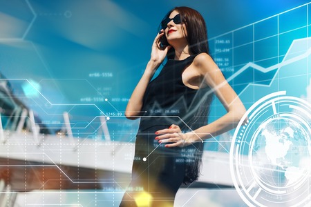 Attractive smiling brunette business woman with red lips wearing elegant black dress and sunglasses standing in front of hi-tech glass building of business center talking on her mobile  phone discussing new project with clients. Financial infographics.の写真素材