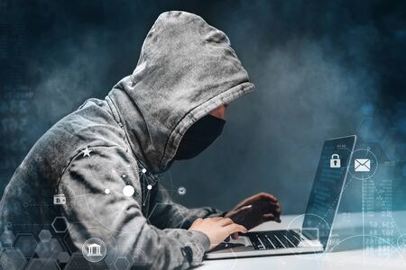 Male hacker wearing hoodie and mask working on a laptop in the dark room trying to get access to personal data. Cyber crime concept.の写真素材
