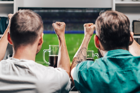 Two male buddies watching live football game broadcast on tv, cheering and drinking beer. Friends spending their day-off together at home.の写真素材