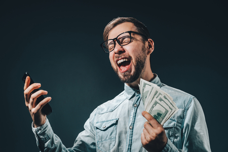 Lucky guy celebrating victory after making bets using gambling mobile application on his mobile phone. Casually dressed bearded business man holding ward of cash in hands.の写真素材