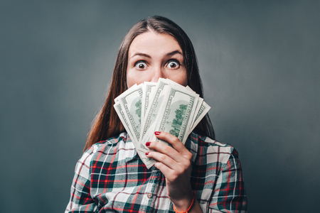 Young attractive casually dressed woman looking shocked holding dollar banknotes in hand.の写真素材